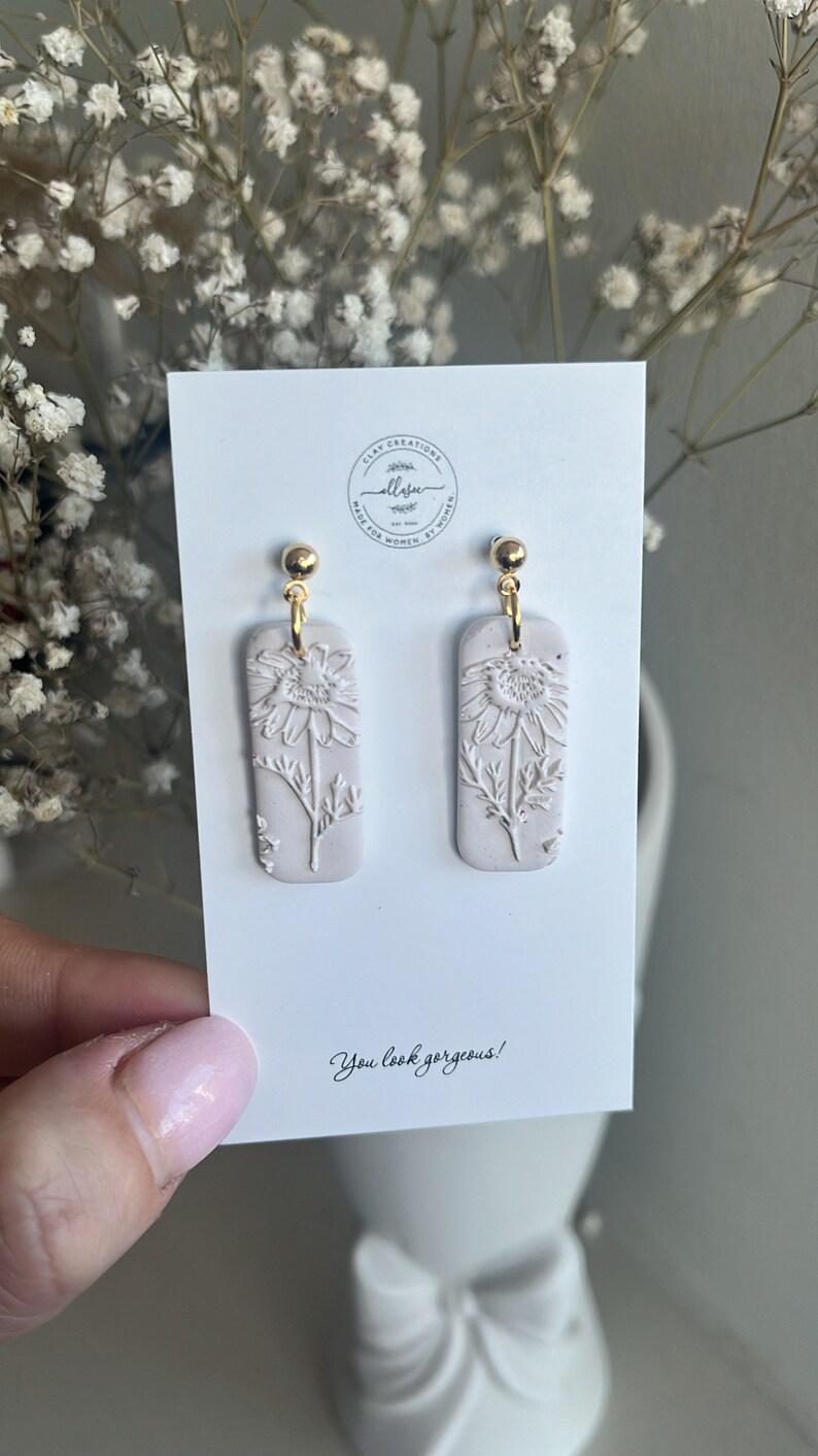 May include: A pair of white clay earrings with a floral design. The earrings are rectangular in shape and have a gold hook.