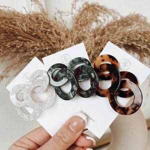 May include: A hand holds several pairs of linked oval hair clips in various colors. The clips are white, dark green, and tortoiseshell. The background includes dried plants and a white surface.
