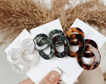 May include: A hand holds several pairs of linked oval hair clips in various colors. The clips are white, dark green, and tortoiseshell. The background includes dried plants and a white surface.