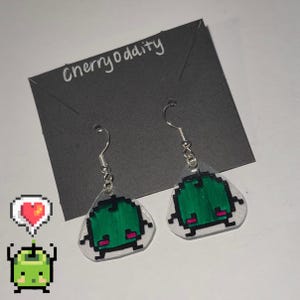 May include: A pair of green pixelated creature earrings with pink accents. The earrings are on a black card with the text "cherryoddity" written in white.