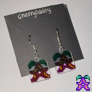 May include: A pair of silver dangle earrings with clear plastic charms featuring a pixelated image of a purple and green plant. The earrings are on a black card with the text "cherryoddity" written in white.