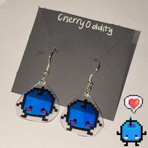 May include: A pair of blue pixelated character earrings with silver hooks. The earrings are made of clear plastic and have a blue pixelated character with pink cheeks on each earring. The earrings are on a black card with the text "cherryo ditty" written in white.