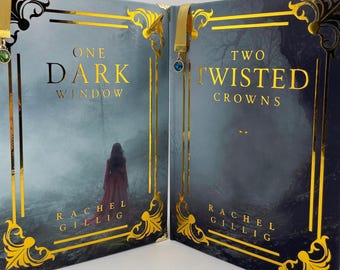 May include: Two hardcover books titled One Dark Window and Two Twisted Crowns by Rachel Gillig. The covers feature dark, misty forest scenes with gold foil filigree frames and metallic bookmarks with small green and blue circular charms attached.