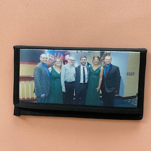 May include: A black wallet with a zipper closure and a printed image of a group of people in formal attire. The image shows six people, three women and three men, standing in front of a staircase.