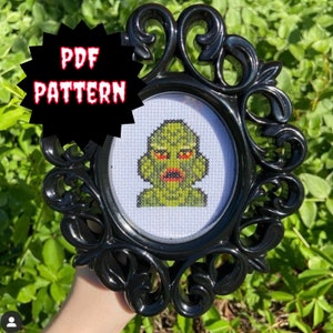 May include: A black framed embroidery hoop with a white cross-stitch design of a green creature with red eyes. The text "PDF PATTERN" is on a black circle.