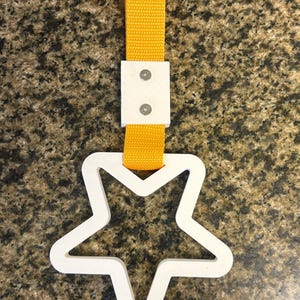 May include: A white star-shaped car accessory with a yellow strap. The star is connected to the strap by a white rectangular piece secured with two screws. The strap is made of a woven material.