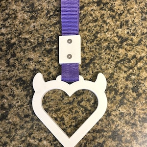 Può includere: Accessorio per auto a forma di cuore bianco con corna di diavolo, attaccato a un cinturino viola. Il cinturino è collegato a un pezzo rettangolare bianco con due rivetti argentati. Progettato per essere appeso all'interno di un'auto.
