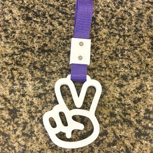May include: A white plastic hand gesture charm in the shape of a peace sign attached to a purple nylon strap. The strap is secured by a white rectangular plastic connector piece featuring two small silver-colored metal screws on the front surface.