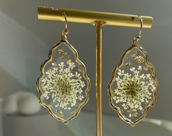 Puede incluir: Un par de pendientes de tono dorado con un diseño de resina transparente que presenta una flor blanca prensada en su interior. Los pendientes están colgados de un soporte dorado.