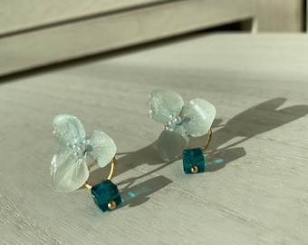 May include: A pair of delicate earrings featuring light blue flower-shaped pendants with small white beads. Each earring has a gold hoop and a teal cube with a gold bead accent. The earrings are displayed on a white wooden surface.