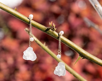 Op de afbeelding: Een paar delicate witte bloemeoorringen met parel accenten die aan een tak hangen. De oorringen zijn gemaakt van een doorschijnend materiaal en hebben een delicate, vrouwelijke uitstraling.