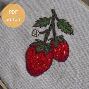 Peut inclure: Art de broderie sur un cercle représentant deux fraises rouges avec des feuilles vertes et une fleur blanche sur un fond en tissu beige. Le texte "PDF pattern" est écrit sur un cercle jaune en haut à gauche.