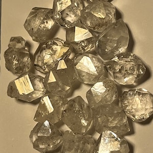 Puede incluir: Una colección de 20 diamantes en bruto, transparentes, facetados y octaédricos. Los diamantes son de varios tamaños y formas, y están dispuestos en una pila sobre una superficie blanca.