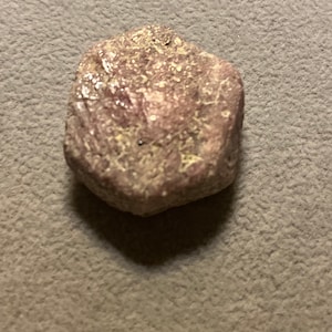 Könnte beinhalten: Ein rauer, unregelmäßig geformter, rosa-violetter Stein mit strukturierter Oberfläche. Der Stein hat kleine, hellfarbige Flecken und ist vor einem grauen Hintergrund platziert.