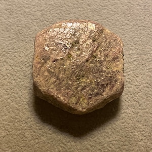 Puede incluir: Una gema en bruto, sin cortar, con una superficie multifacética. La piedra tiene un tono púrpura apagado con toques de verde y un aspecto texturizado. La gema tiene forma irregular.