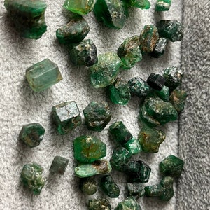 May include: A collection of rough, green emerald crystals of various sizes and shapes. The crystals are scattered on a gray, textured surface.