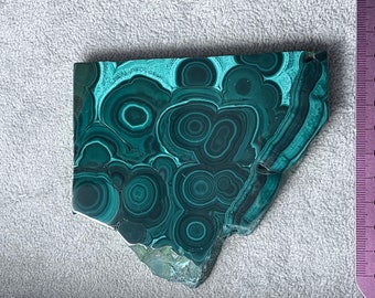 May include: A polished malachite stone slab with a vibrant green and dark green swirling pattern. The stone has a rough, uneven edge and is next to a ruler marked in inches and centimeters.