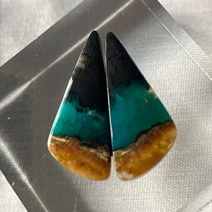 Pode incluir: Um par de brincos de cabochão em forma de lágrima com um esquema de cores preto, turquesa e laranja.