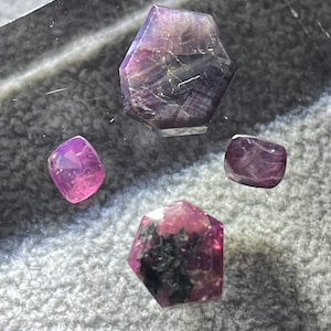 May include: Four faceted purple gemstones of varying sizes. The gemstones are displayed on a clear surface.