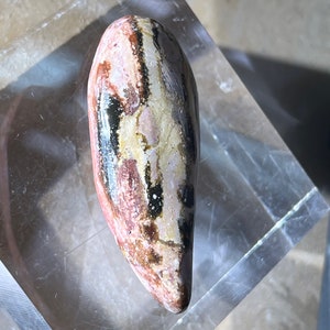 May include: A smooth, polished, elongated stone with a unique pattern of red, white, and black stripes. The stone is displayed on a clear acrylic base.
