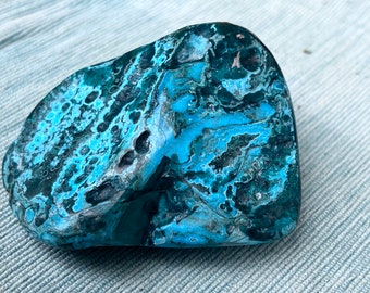 May include: A polished stone with a striking pattern of teal, turquoise, and dark blue hues. The stone's surface displays a complex, swirling design, with areas of lighter and darker coloration. The overall shape is rounded and irregular, suggesting a natural origin.