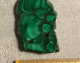 May include: A green malachite stone with a swirling pattern. The stone is about 2 inches long and 1 inch wide. It is sitting on a ruler with markings in inches and centimeters.