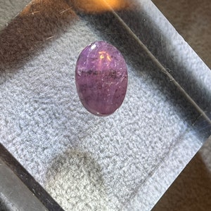 May include: A purple oval-shaped gemstone with a sparkly, iridescent finish. The gemstone is displayed on a clear acrylic block.