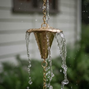 May include: A brass rain chain with water cascading from the decorative cone-shaped top. The chain is suspended, with water flowing down the links. The background is blurred, with green foliage and a white house visible.
