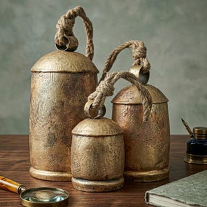 Puede incluir: Tres campanas decorativas de diferentes tamaños con acabado dorado envejecido y asas de cuerda natural. Están dispuestas sobre una superficie de madera, evocando un estilo vintage o rústico. El conjunto destaca las texturas y los materiales naturales.