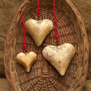 May include: Three gold-coloured heart-shaped ornaments with a distressed finish, suspended from red cords. The hearts are arranged in a woven basket, creating a rustic display. The hearts vary in size, adding visual interest.