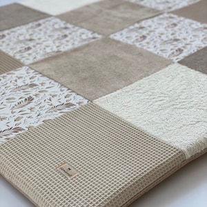 Könnte beinhalten: Eine beige und weiße Patchwork-Spielmatte mit einer Waffel-Oberfläche. Die Matte hat eine Vielzahl von Texturen und Mustern, darunter einen Blumendruck, eine Waffelstruktur und einen weichen, plüschigen Stoff. Die Matte ist perfekt für Babys und Kleinkinder zum Spielen und Entdecken.