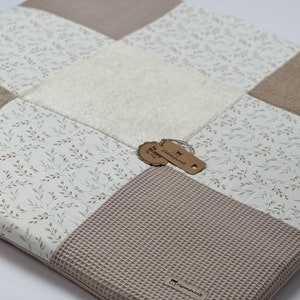 Op de afbeelding: Een beige en witte patchwork baby speelmat met een wit, pluche vierkant in het midden. De mat heeft een patroon van kleine bruine takken op een witte achtergrond. De mat heeft twee bruine labels met de tekst "Kleinkindwelt" erop.
