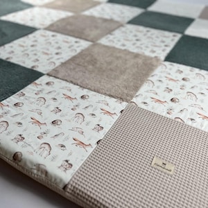 Puede incluir: Una alfombra de juego de patchwork con una variedad de texturas y patrones. La alfombra presenta un esquema de color verde, marrón y blanco con un estampado de animales del bosque. La alfombra está hecha de materiales suaves y cómodos y es perfecta para bebés y niños pequeños.