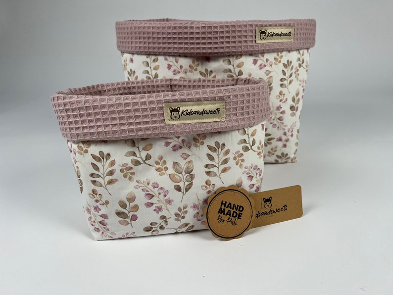 May include: Two pink and white fabric storage baskets with a floral pattern. The baskets have a pink waffle-textured top and a brown label with the text "kidsandweets". A brown tag with the text "HAND MADE by Kati" is in the foreground.