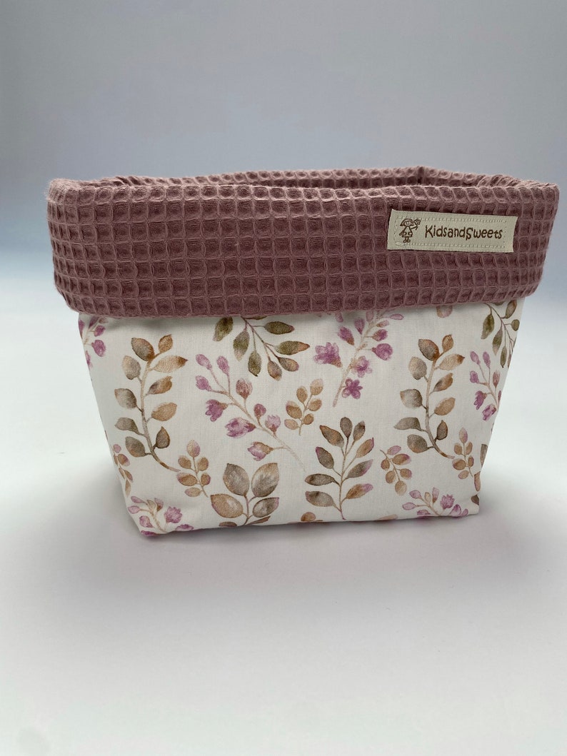 May include: A pink and white fabric storage basket with a floral pattern and a waffle-textured top. The basket has a label that says "KidsandSweets".