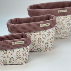 May include: Three pink and white fabric storage baskets with a floral pattern and the text "KidsandQueens" on the front. The baskets are nested inside each other, with the largest basket at the top and the smallest basket at the bottom.