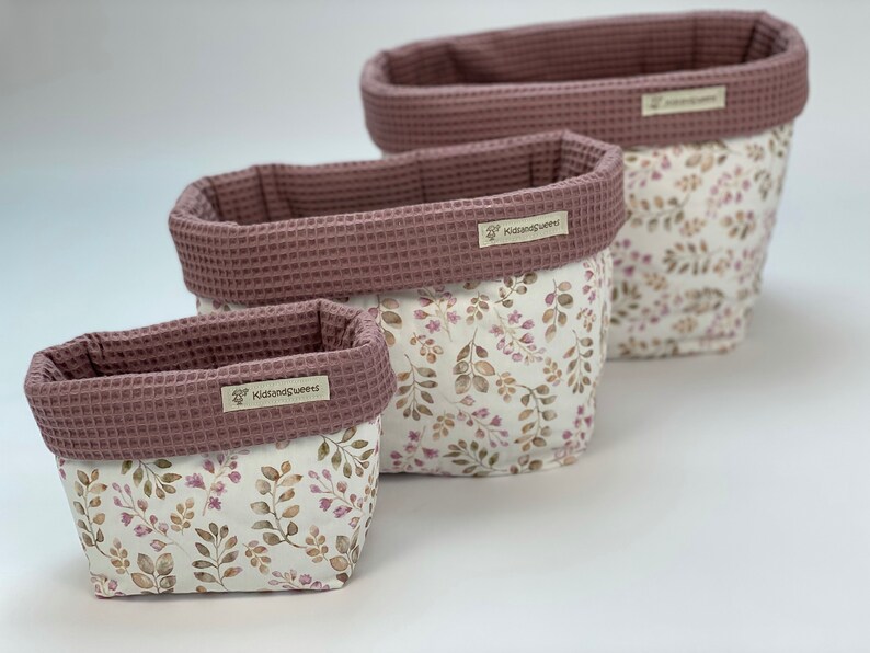 May include: Three pink and white fabric storage baskets with a floral pattern and the text "KidsandQueens" on the front. The baskets are nested inside each other, with the largest basket at the top and the smallest basket at the bottom.