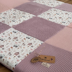 Könnte beinhalten: Ein Patchwork-Quilt in Rosa- und Lilatönen. Der Quilt besteht aus Quadraten mit unterschiedlichen Texturen und Mustern, darunter ein Bauernhof-Tierdruck, Waffelstrick und Plüschstoff. Ein braunes Etikett mit "Hand Made" ist angebracht.
