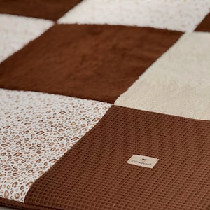 Peut inclure: Gros plan sur une couverture patchwork avec une variété de textures et de motifs. La couverture présente des carrés de tissu marron, crème et imprimé léopard. Une petite étiquette est cousue sur la couverture.