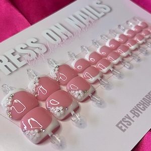 May include: A set of 10 pink and white press-on nails with white tips and small white embellishments. The nails are displayed on a white card with the text "PRESS ON NAILS" and "ETSY-BYCHARRELLIZA".
