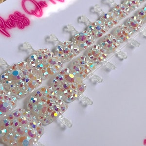 May include: A set of white press-on nails with iridescent rhinestones. The nails are arranged on a white card with the text "Press On Nails" in pink neon lettering and "Oh By Charlizebeth" in pink lettering.