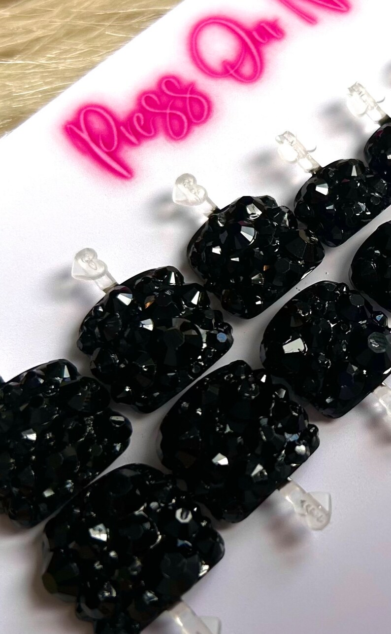 May include: Set of ten black press-on nails with clear adhesive tabs. Each nail is covered in black rhinestones.