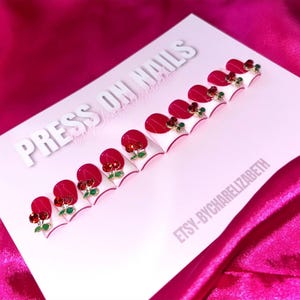 May include: A set of press-on nails with a cherry design. Each nail features a red cherry accent with red gems and green leaves on a white and red base. The words "PRESS ON NAILS" are visible.