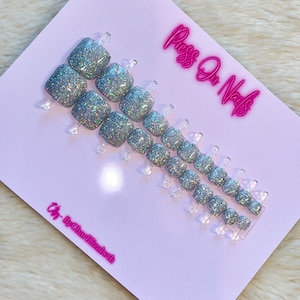 May include: A set of silver glitter press-on nails with a square shape. The nails are arranged in two rows on a white card with the text "Press On Nails" and "Cig - By Charlizabuth" printed in pink.