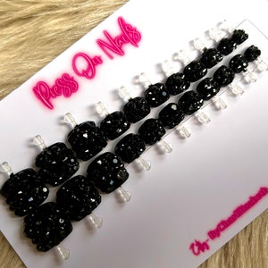 May include: Press-on nails with a black base and a full coverage of black rhinestones. The nails are displayed on a white card with the text "Press On Nails" in pink.