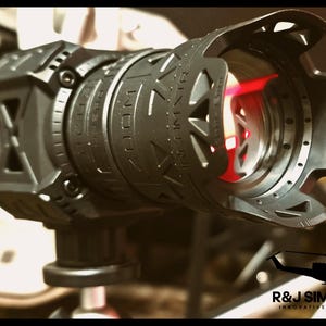 May include: A black camera lens accessory featuring a geometric cutout design and a protective hood. The barrel is marked with the text ZOOM and INFINITY. A bright red light reflects within the lens glass. The R&J SIMTECH INNOVATIVE DESIGNS logo is visible.