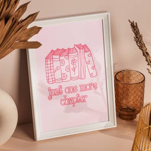 May include: A white framed print with a pink background featuring a stack of books with a star, flowers, and hearts on the covers. The text "just one more chapter" is written in pink below the books.