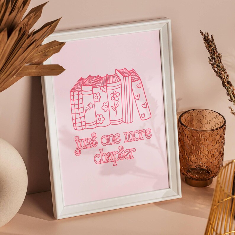 May include: A white framed print with a pink background featuring a stack of books with a star, flowers, and hearts on the covers. The text "just one more chapter" is written in pink below the books.