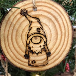May include: A wooden Christmas ornament with a wood-burned design of a gnome wearing a hat. The ornament is round and has a natural wood finish.