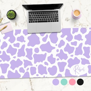 May include: A purple and white cow print desk mat with the word "Rebel" written in cursive on the bottom right corner. The mat is on a white marble surface with a laptop, a potted plant, a cup of coffee, and a pair of glasses.